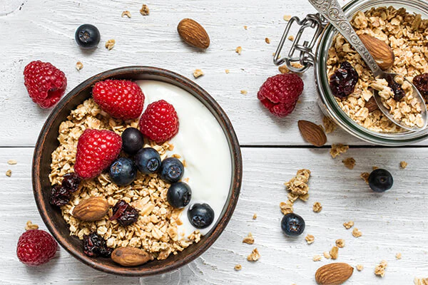 Image d'un bol contenant des graines, des fruits secs et des fruits frais dans du lait et entourés de flocons d'avoine, de framboises, d'amandes et de myrtilles.